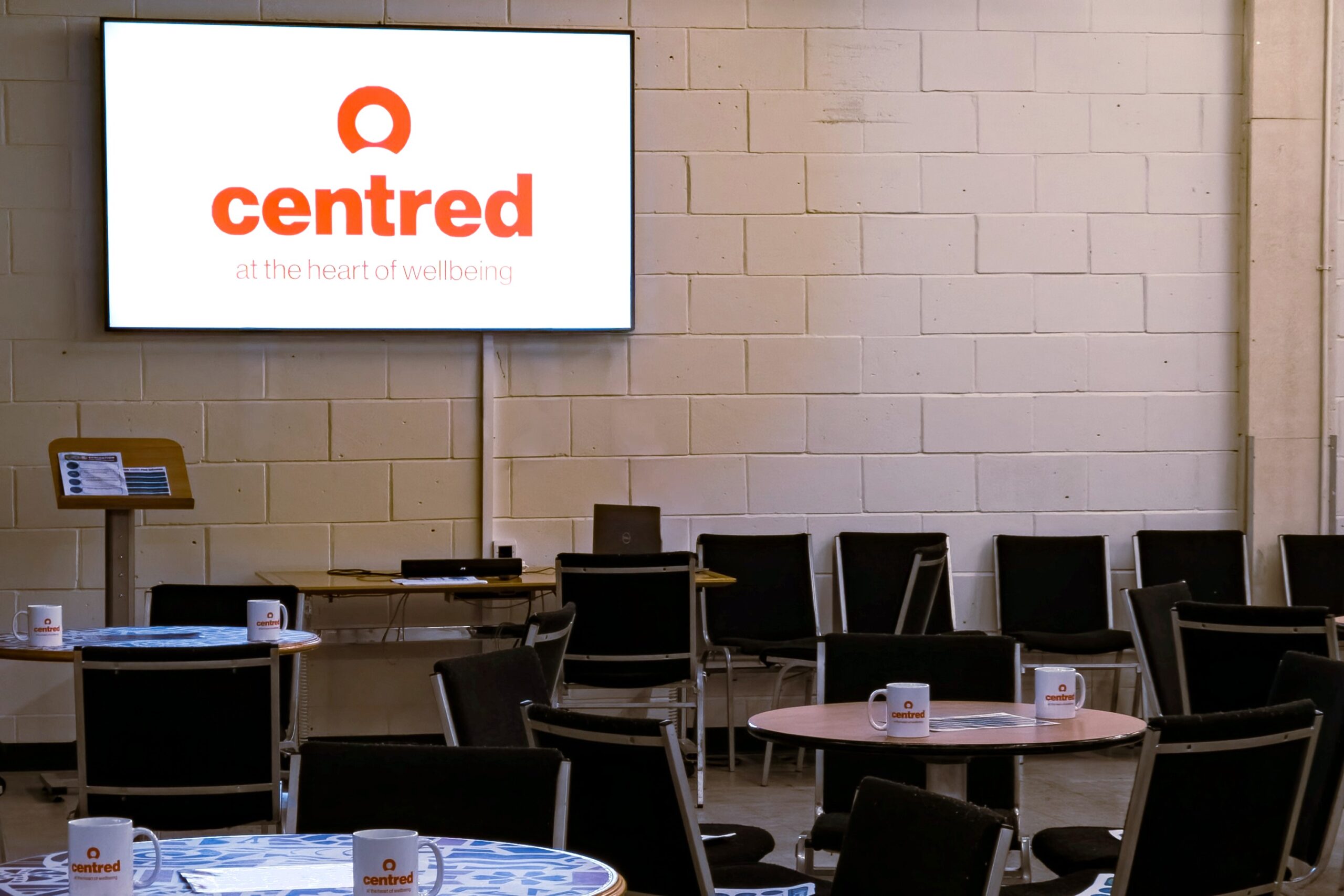 Room with tables and chairs and a large screen on the wall.