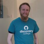 A man with red hair and a beard wearing a Discovery College t-shirt and smiling