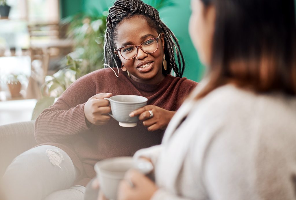 Two women with hot cups talking on a sofa together