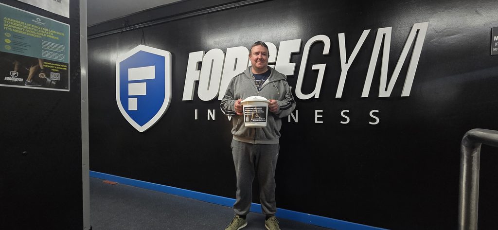 A man in front of a gym sign wearing casual clothes and holding a donation bucket