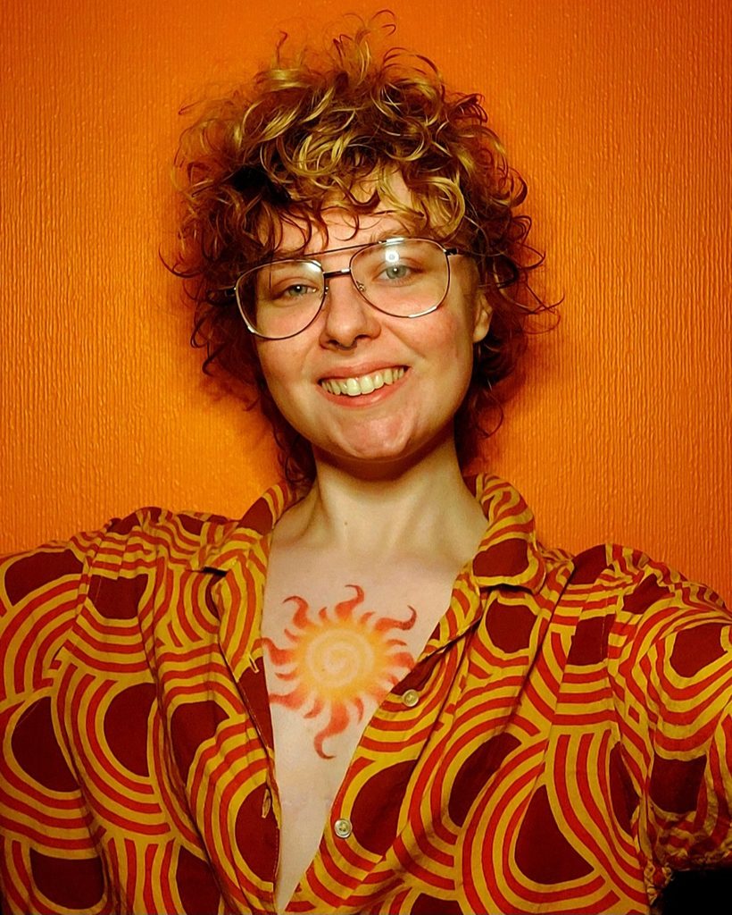 Photo of an artist with curly hair and glasses, smiling at the camera.